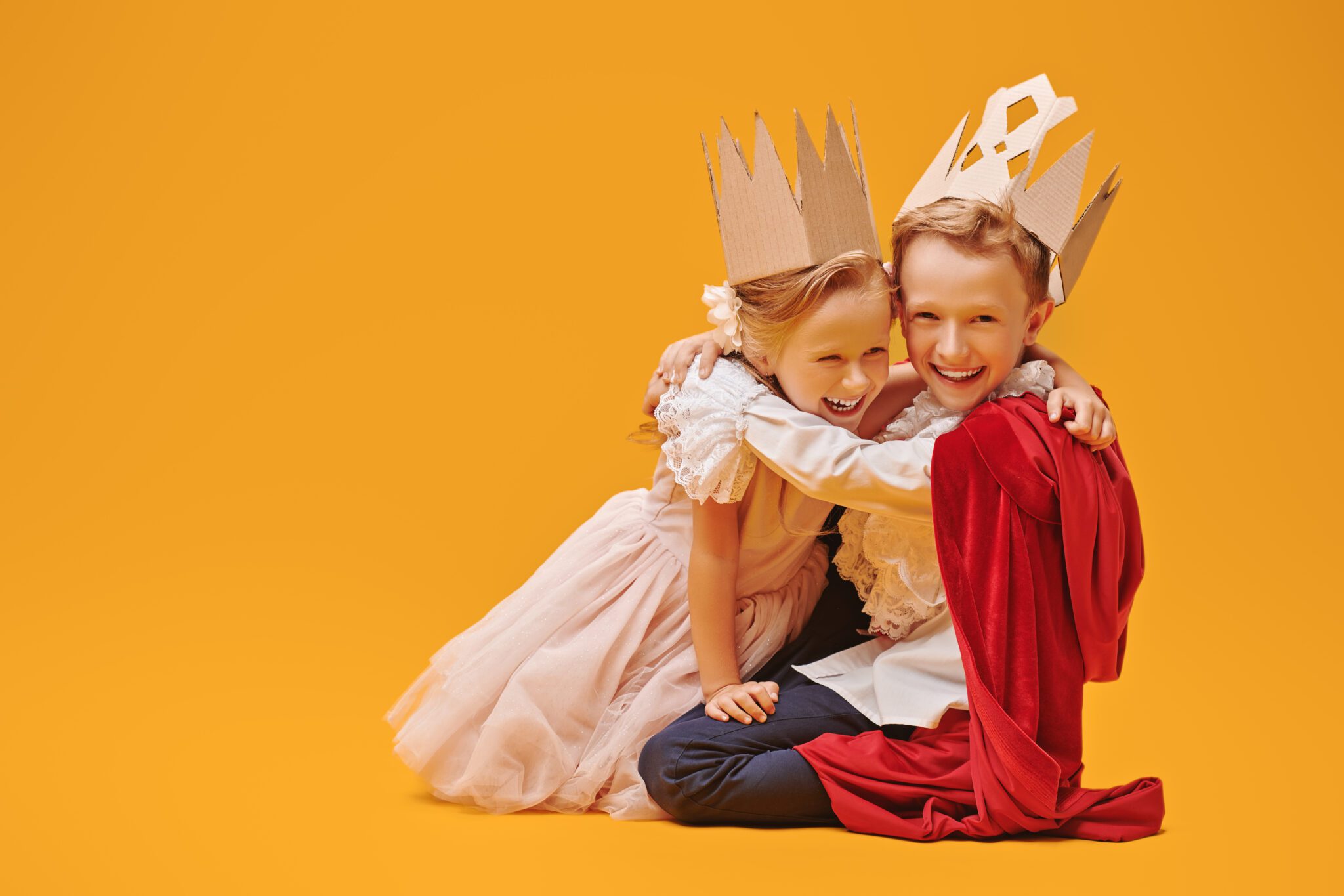 A boy and a girl in Prince and Princess costumes laugh and hug. Childhood dreams. Fantasy, imagination. Studio portrait on a yellow background.