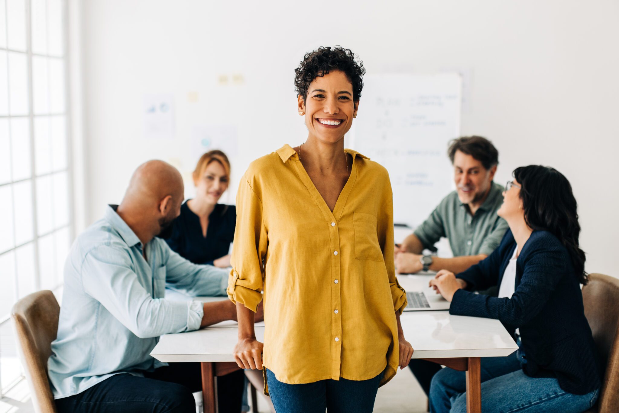 Black business woman standing in a boardroom with her colleagues discussing in the background. Happy female professional working in a marketing agency. Team meeting in a creative office.